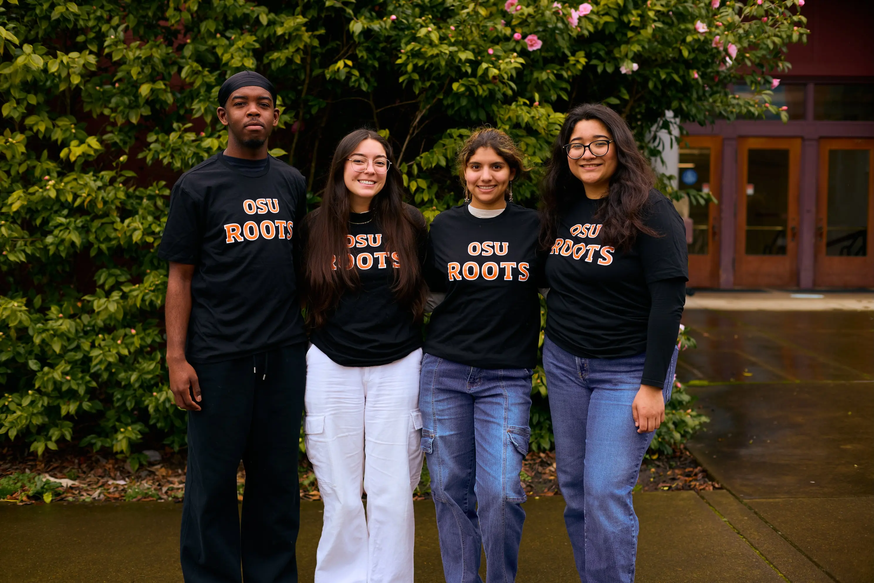 Four students wearing Roots shirts pose in front of green shrubs.